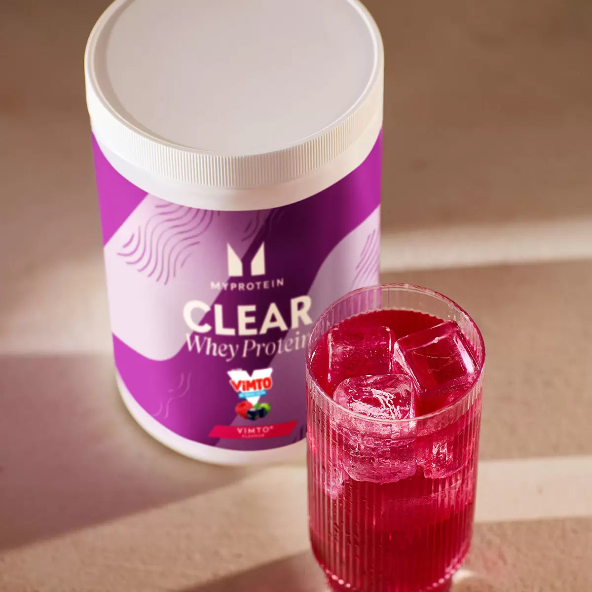 Glass of clear, purple Myprotein Vimto protein drink next to the powder tub.