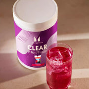 Glass of clear, purple Myprotein Vimto protein drink next to the powder tub.