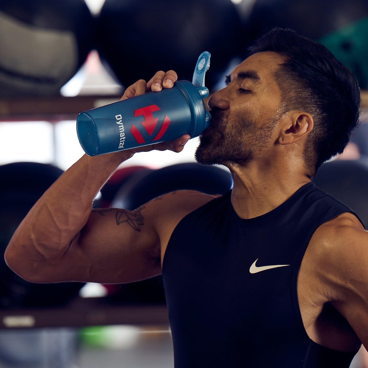 Man drinking from a blue Dymatize protein shaker in a gym setting