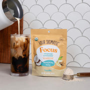 Four Sigmatic Focus Organic Creamer package with a glass of iced coffee and a scoop of creamer on a neutral background.