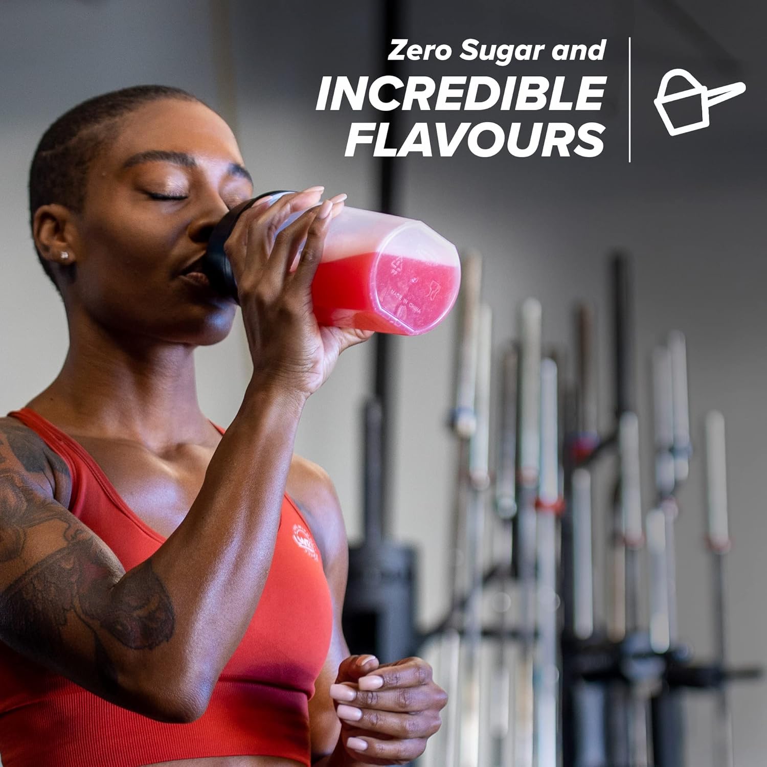 Woman in red sports bra drinking a pink supplement shake in a gym setting with text about zero sugar and incredible flavors.