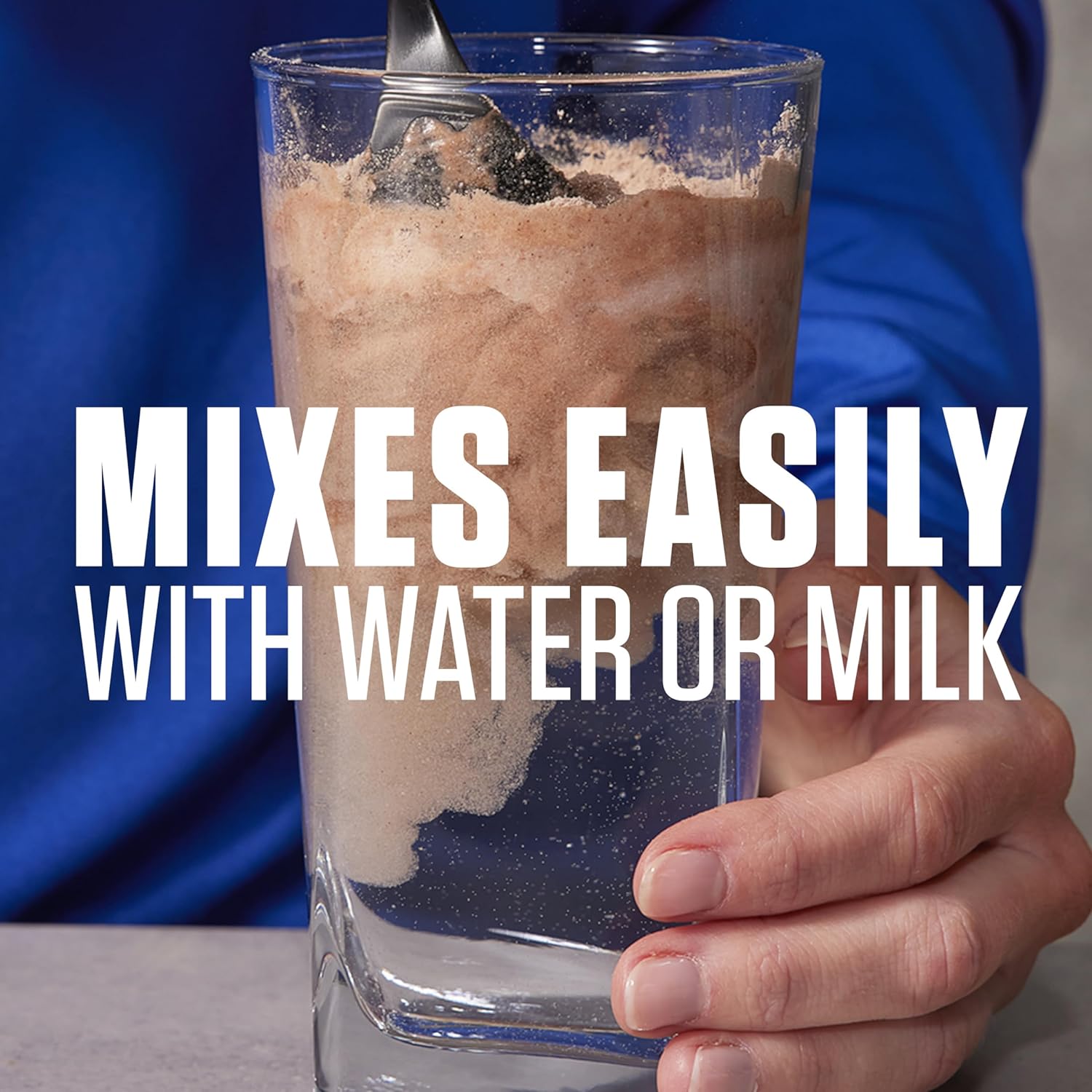 Glass of a brown liquid with a spoon, held by a hand, against a blue background with text 'Mixes Easily with Water or Milk'.
