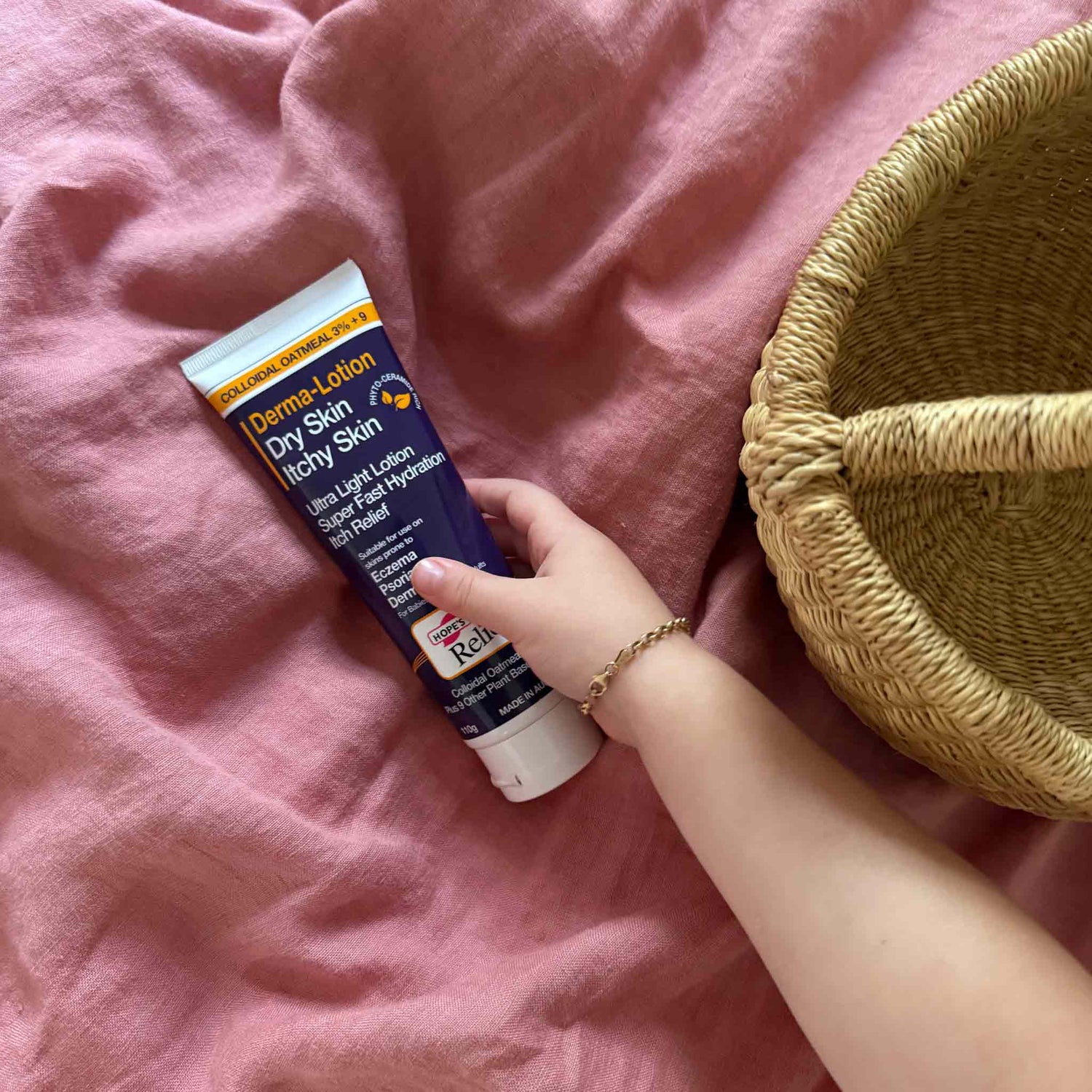 Hand holding a tube of Derma-Lotion on a pink surface with a woven basket.