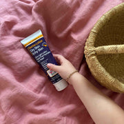 Hand holding a tube of Derma-Lotion on a pink surface with a woven basket.