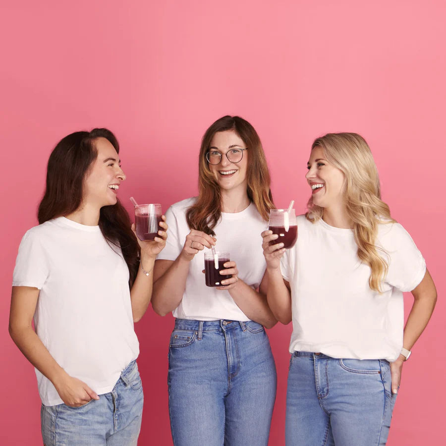 Three women in white t-shirts and jeans holding drinks against a pink background