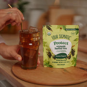 Person holding a glass of iced tea next to a package of Four Sigmatic Protect organic herbal tea on a wooden surface.