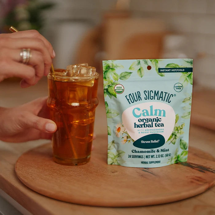 Person holding a glass of iced tea next to a package of Four Sigmatic Calm organic herbal tea on a wooden surface.