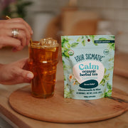Person holding a glass of iced tea next to a package of Four Sigmatic Calm organic herbal tea on a wooden surface.
