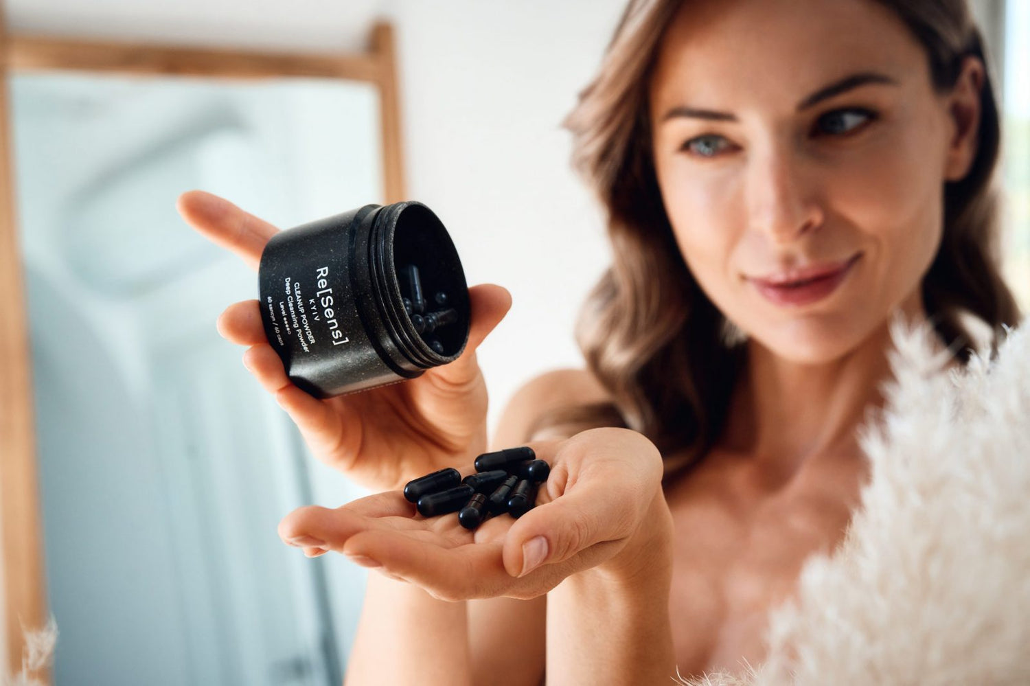 Woman holding a container of black powder and some black capsules in her hand.