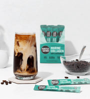 Glass of iced coffee with Hunter Gather Marine Collagen Unflavored packets and a bowl of chocolate chips on a white background.